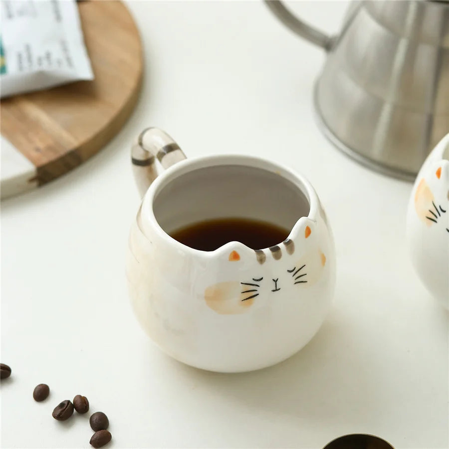 White ceramic cat-shaped coffee mug with striped tail handle, filled with black coffee, surrounded by coffee beans and kettle.