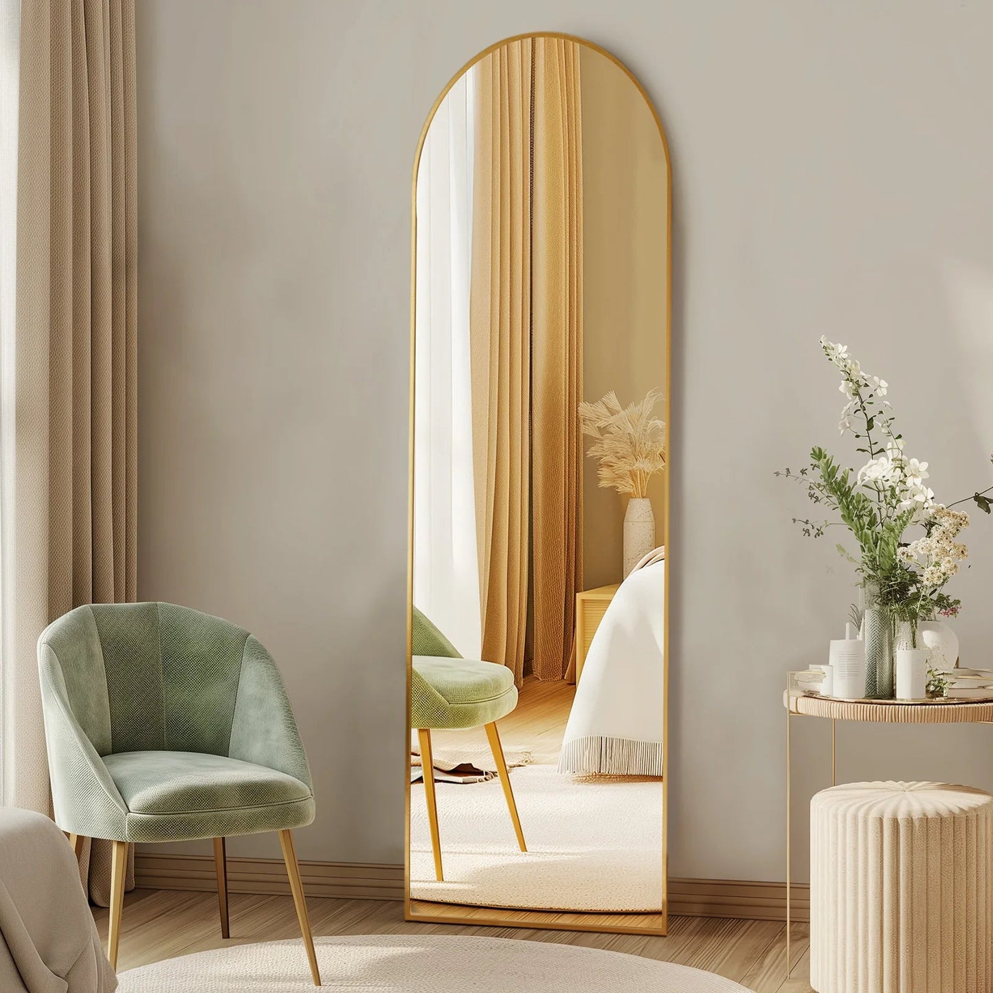 Arched full length standing mirror with gold metal frame in modern bedroom with green chair and neutral decor