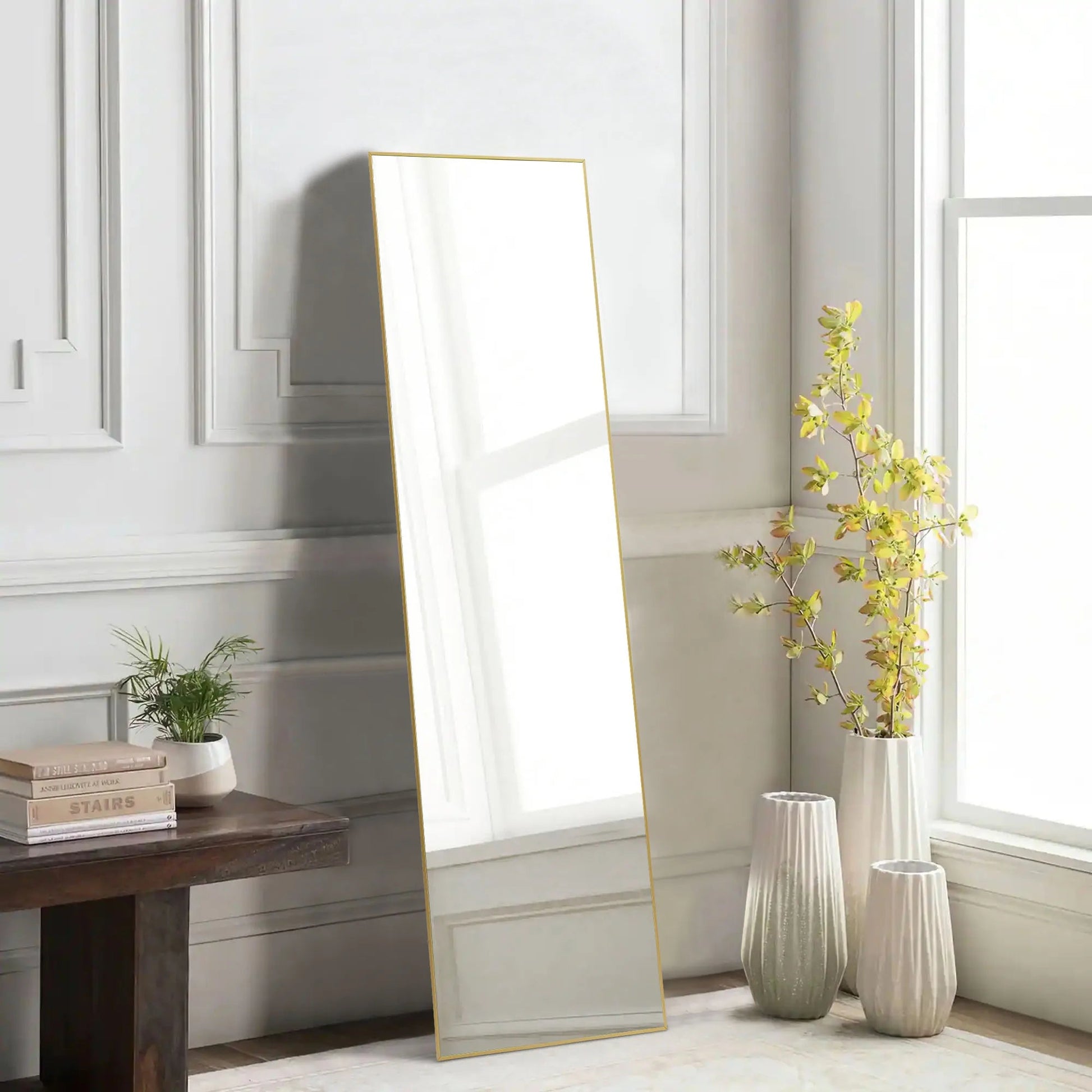 Full length rectangular mirror with thin gold metal frame leaning against white paneled wall beside vases and plant