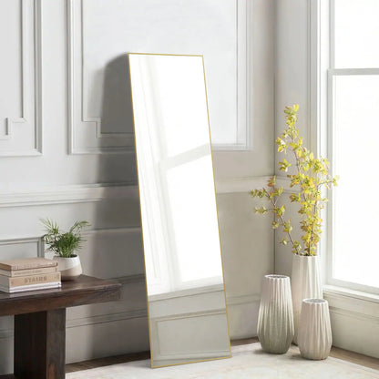 Full length rectangular mirror with thin gold metal frame leaning against white paneled wall beside vases and plant