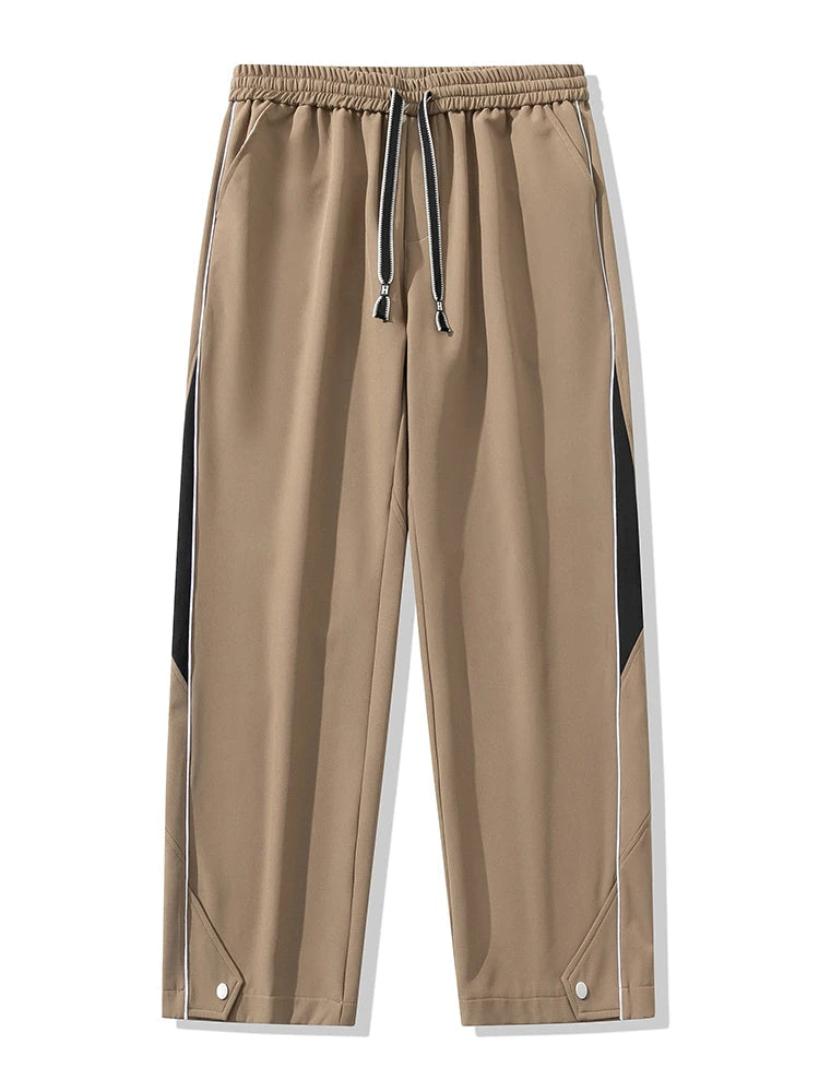 Men's khaki wide-leg track pants with black side panels, white piping, drawstring waist, and snap button hem details.