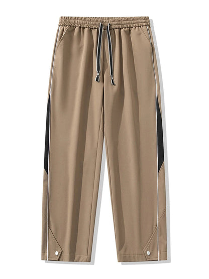 Men's khaki wide-leg track pants with black side panels, white piping, drawstring waist, and snap button hem details.
