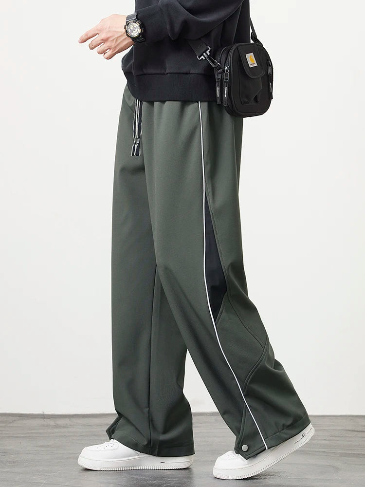 Men's dark green wide-leg track pants with white piping detail and drawstring waist, styled with white sneakers and black crossbody bag