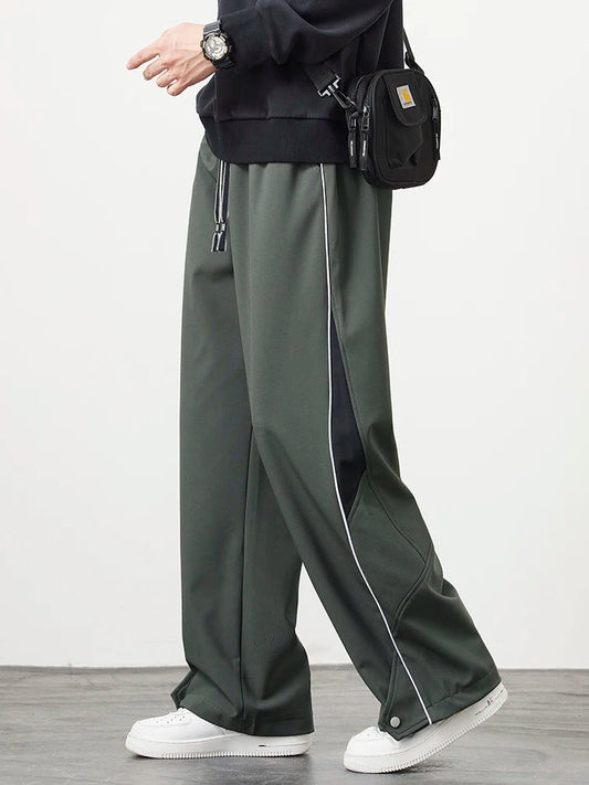Men's dark green wide-leg track pants with white piping detail and drawstring waist, styled with white sneakers and black crossbody bag