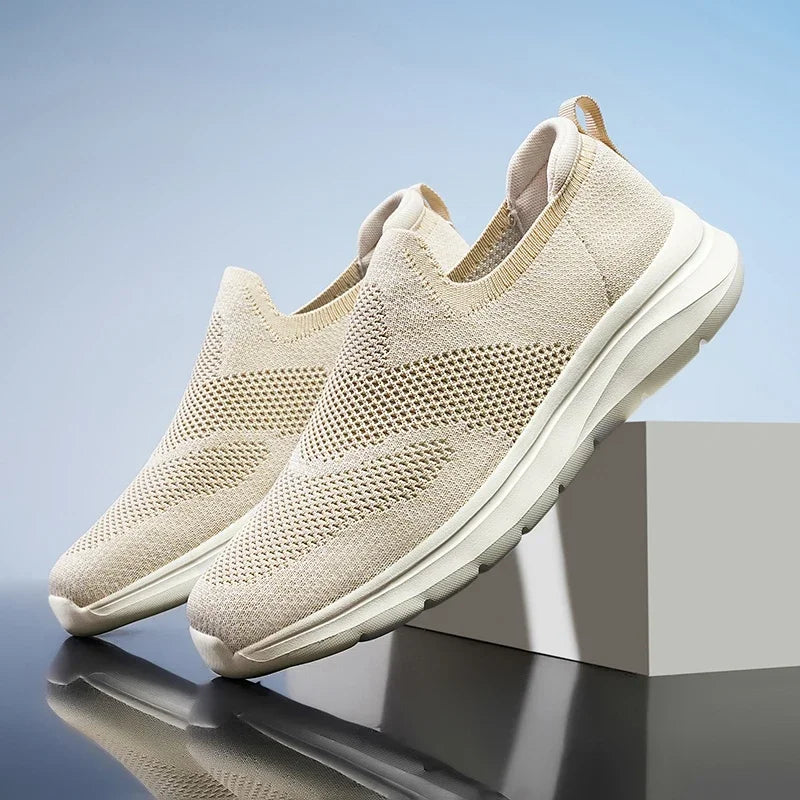 Beige women's slip-on mesh knit sneakers with white sole, displayed on a geometric platform against blue background.