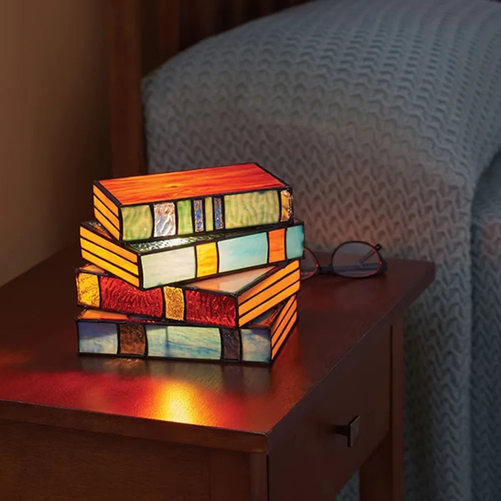 Tiffany-style stained glass lamp shaped like stacked colorful books glowing on a wooden bedside nightstand.