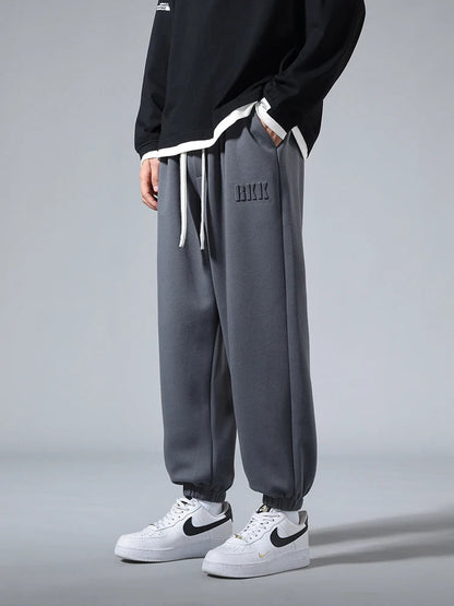 Men's gray tapered knit joggers with drawstring waist, embroidered logo, elastic cuffs, paired with white Nike sneakers.