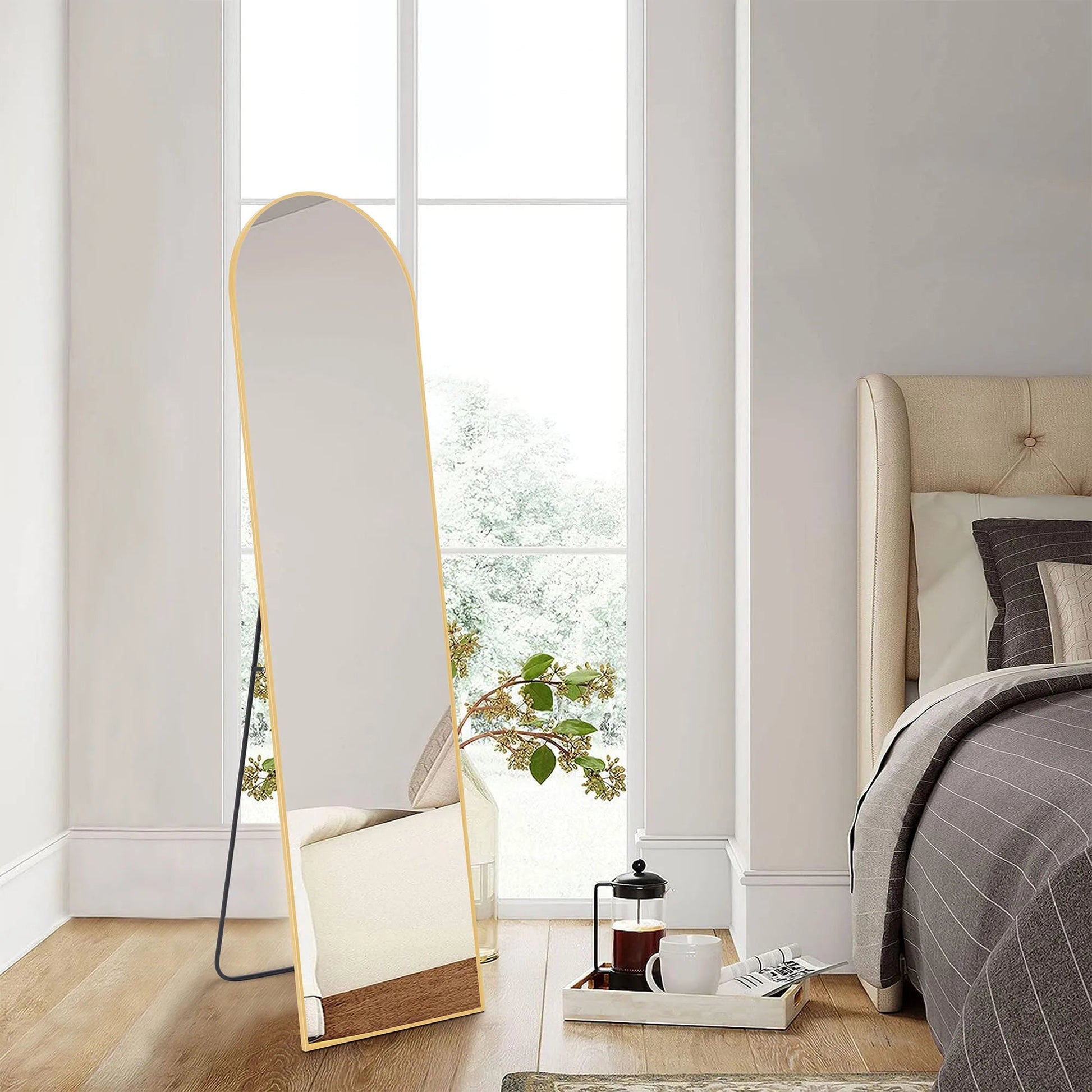 Tall arched full-length floor mirror with thin gold metal frame leaning against white bedroom wall on wood floor.