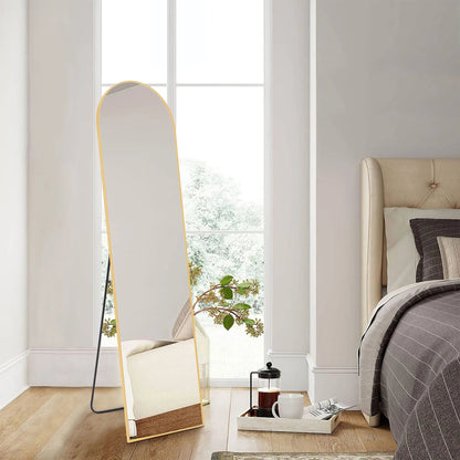 Tall arched full-length floor mirror with thin gold metal frame leaning against white bedroom wall on wood floor.