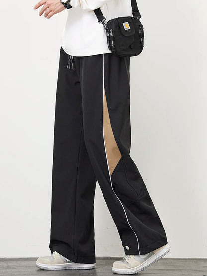 Men's black wide-leg track pants with tan contrast panels, white piping, snap hem, worn with white sneakers and crossbody bag.