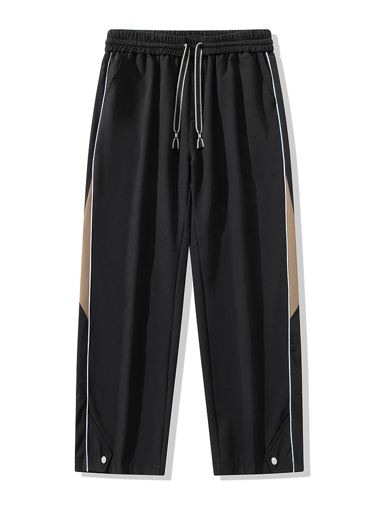 Black wide-leg men's track pants with khaki side panels, white piping, drawstring waist, and snap hem detail.