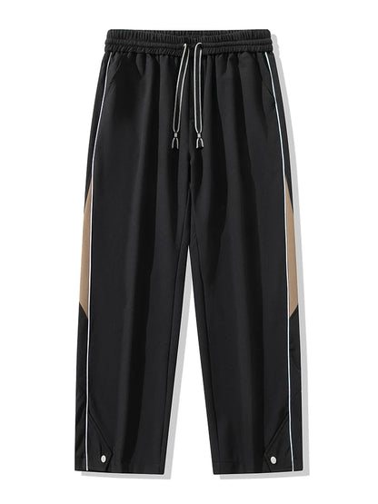 Black wide-leg men's track pants with khaki side panels, white piping, drawstring waist, and snap hem detail.