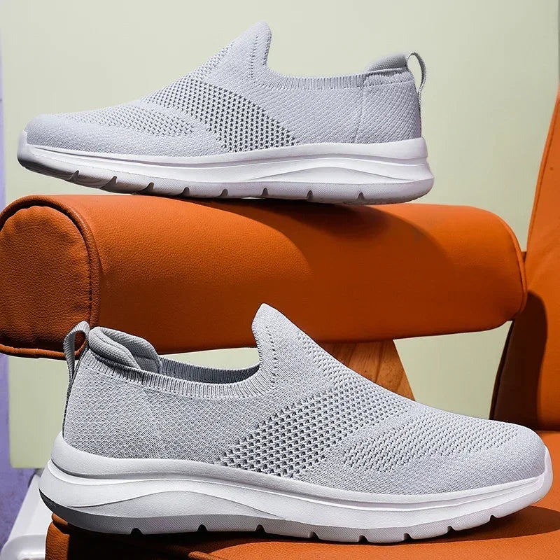 Light gray women's slip-on mesh sneakers with white sole, shown from side and front angles on orange props.