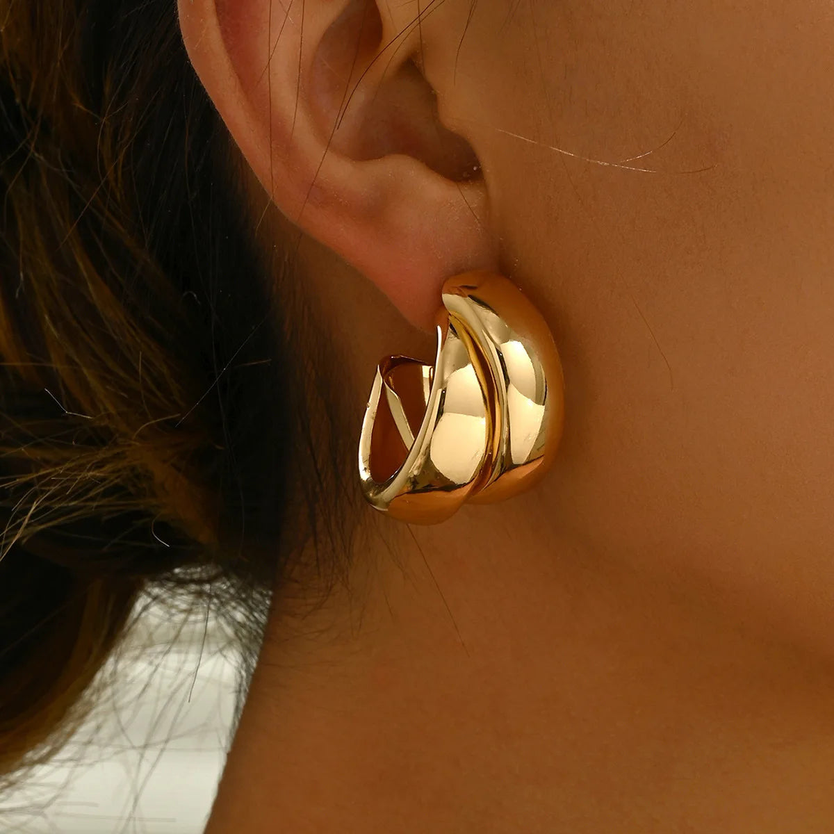 Woman wearing large twisted gold-tone chunky hoop earrings with polished finish and layered crossover design