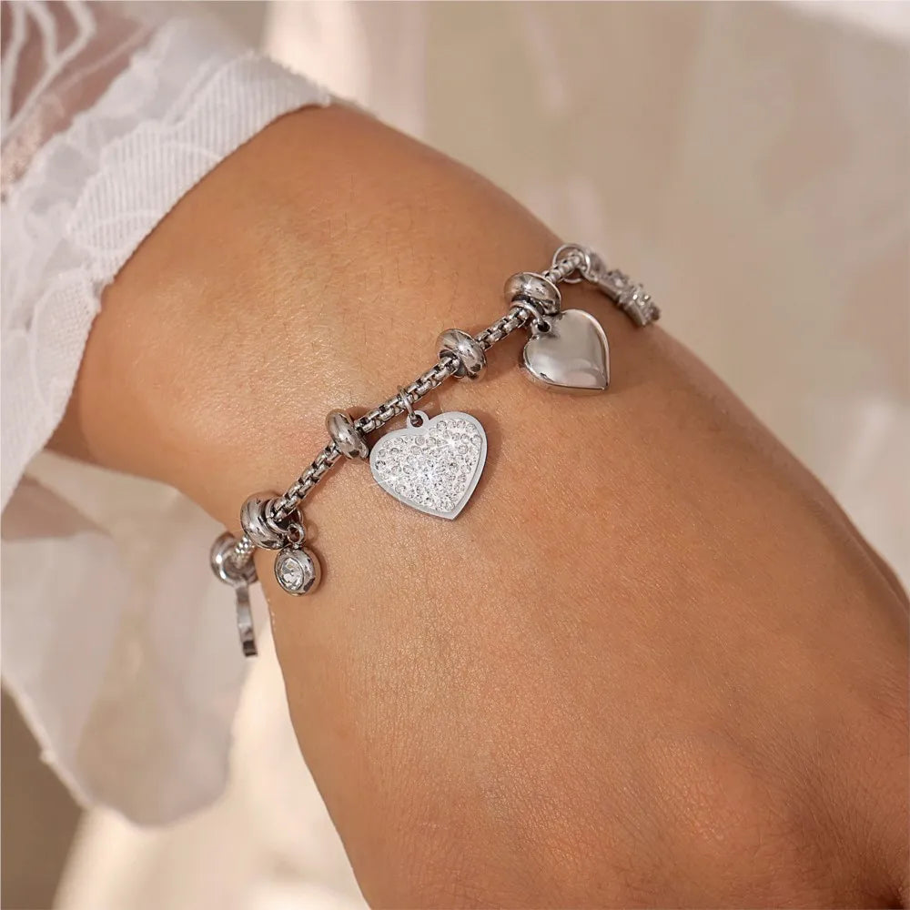 Silver chain bracelet with crystal-studded and polished heart charms worn on a woman's wrist, white lace sleeve background.