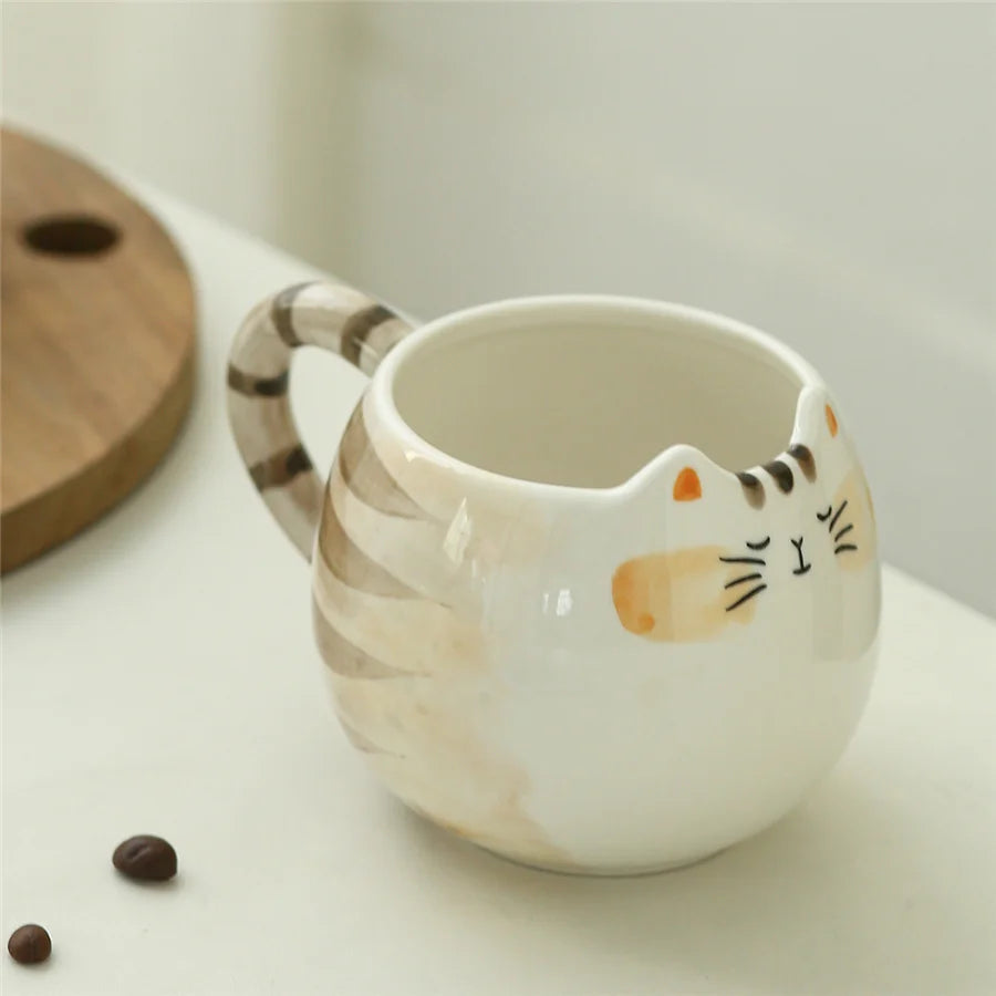 White ceramic cat-shaped coffee mug with striped tail handle, featuring painted cat face, on white surface with wooden lid nearby.