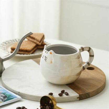 White ceramic cat-shaped coffee mug with striped tail handle, filled with coffee, displayed on marble and wood tray with biscuits nearby.