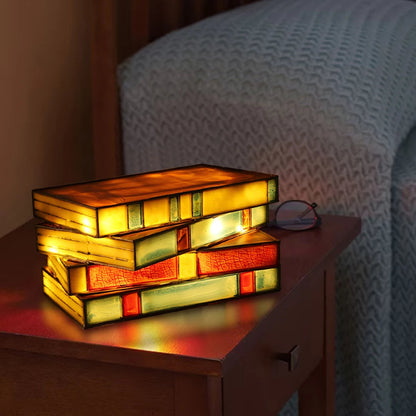 Stained glass lamp shaped like stacked books glowing warmly on a wooden nightstand beside reading glasses.