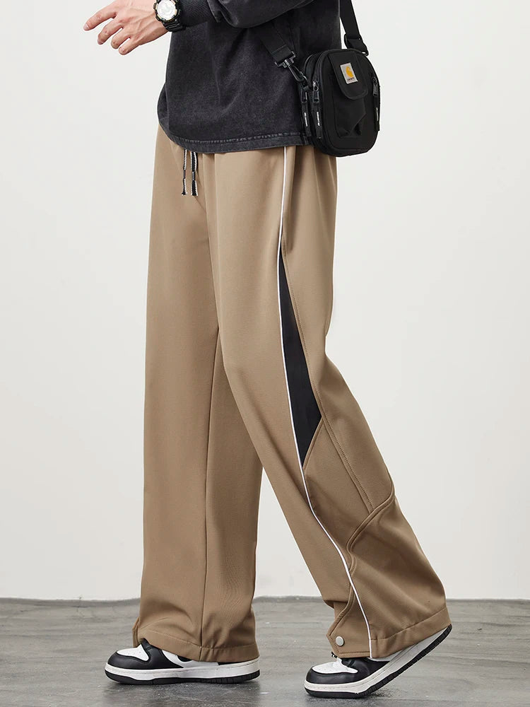 Men's khaki wide-leg track pants with black side piping detail, drawstring waist, and snap hem, styled with sneakers.