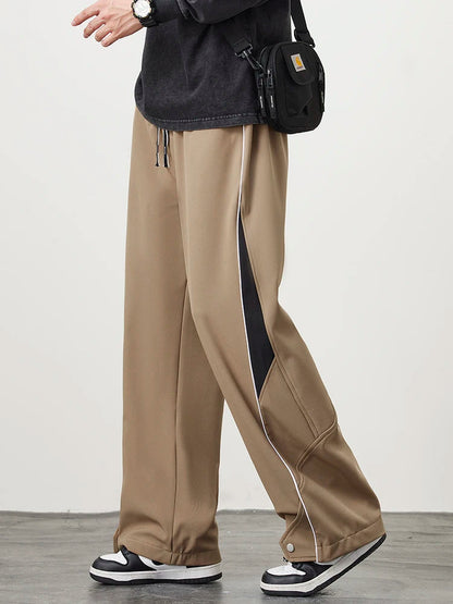 Men's khaki wide-leg track pants with black side piping detail, drawstring waist, and snap hem, styled with sneakers.