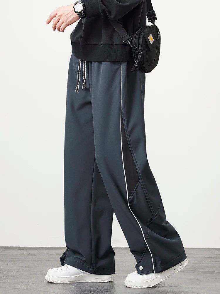 Men's wide-leg track pants in dark gray with white piping, drawstring waist, and snap hem detail, styled with white sneakers.