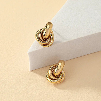 Gold tone interlocked knot hoop stud earrings displayed on white geometric surface against cream background