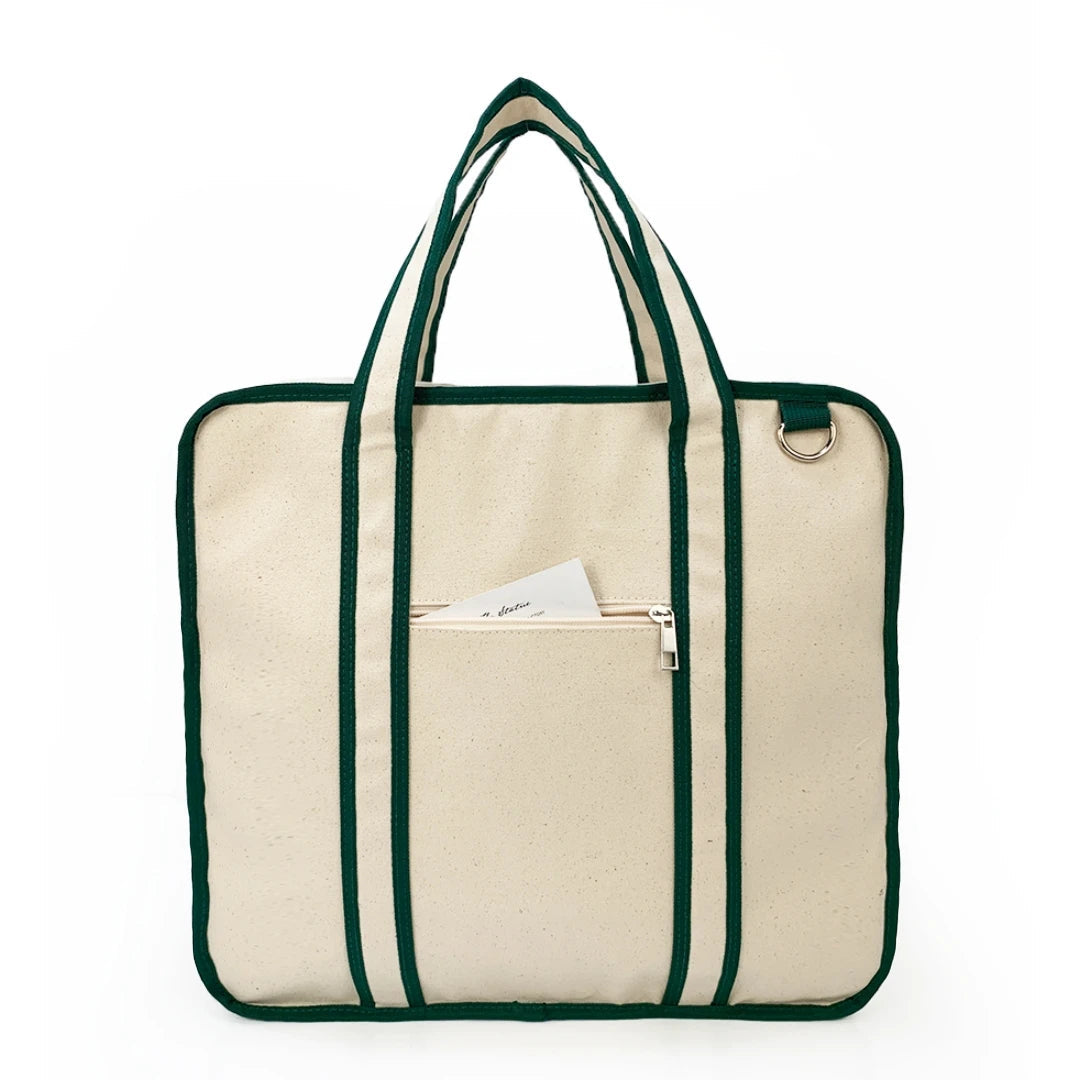 Natural canvas tote bag with dark green trim, dual handles, front zip pocket, and silver D-ring, shown against white background.