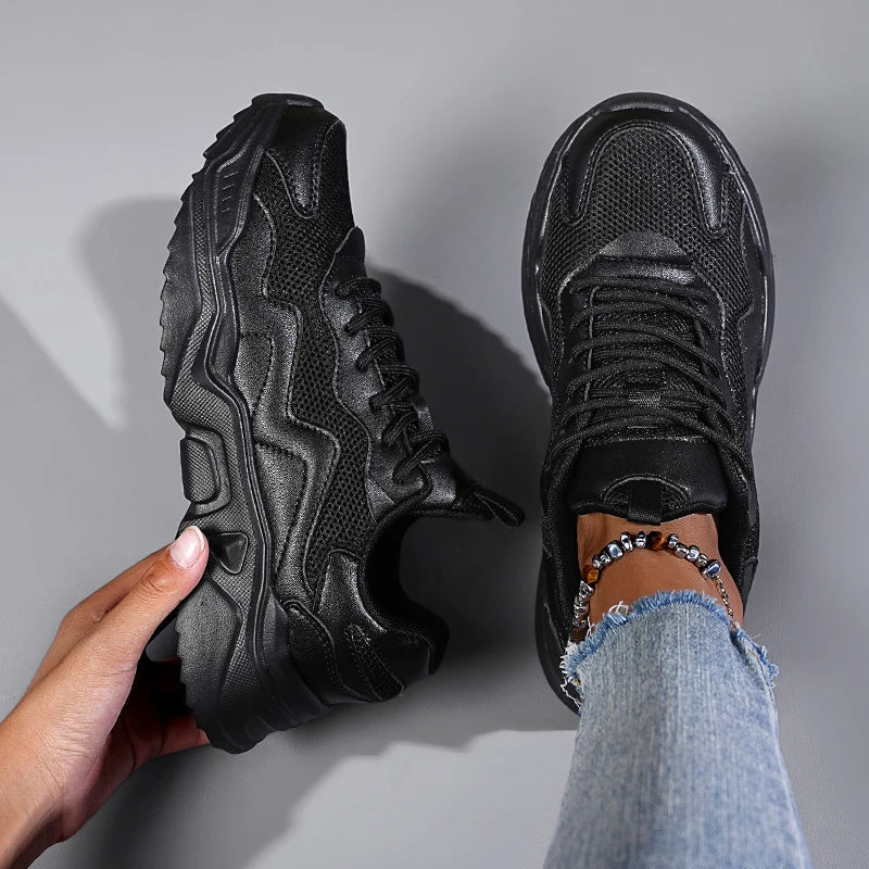 Black women's mesh lace-up chunky sneakers shown from above, one held in hand, other worn with distressed jeans and anklet.