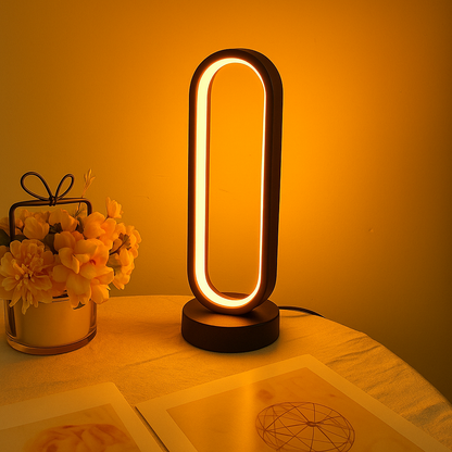 LED oval-shaped desk lamp emitting warm amber light, displayed on a table beside yellow flowers and decorative items.