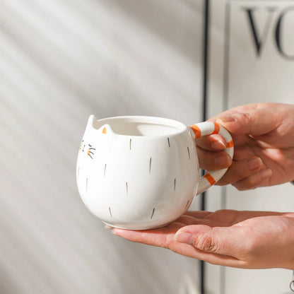 White ceramic cat-shaped coffee mug with painted whiskers, ears, black markings, and orange striped tail handle, held in two hands.