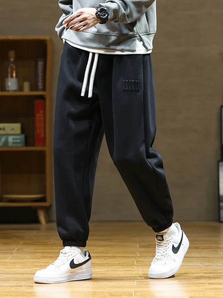 Men's black drawstring sweatpants with embroidered logo, elastic cuffs, worn with gray sweatshirt and white Nike sneakers.