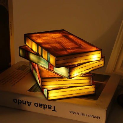 Glowing stained glass stacked books lamp with warm amber light, sitting atop a Tadao Ando book on a shelf.