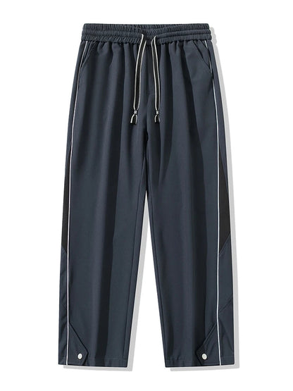 Men's wide-leg track pants in dark gray with black side panels, white piping, drawstring waist, and snap hem details.