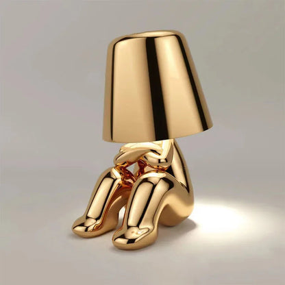 Gold metallic thinker figurine desk lamp with lampshade head, seated pose, emitting warm ambient light on gray background