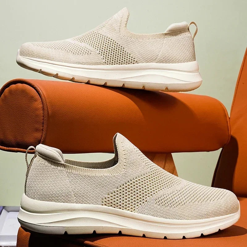 Beige slip-on mesh sneakers with white soles displayed on orange props, shown from side and front angles.