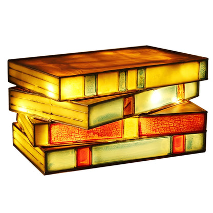 Tiffany-style stained glass lamp shaped like stacked books glowing warm amber, yellow, and red, perfect for desk decor.