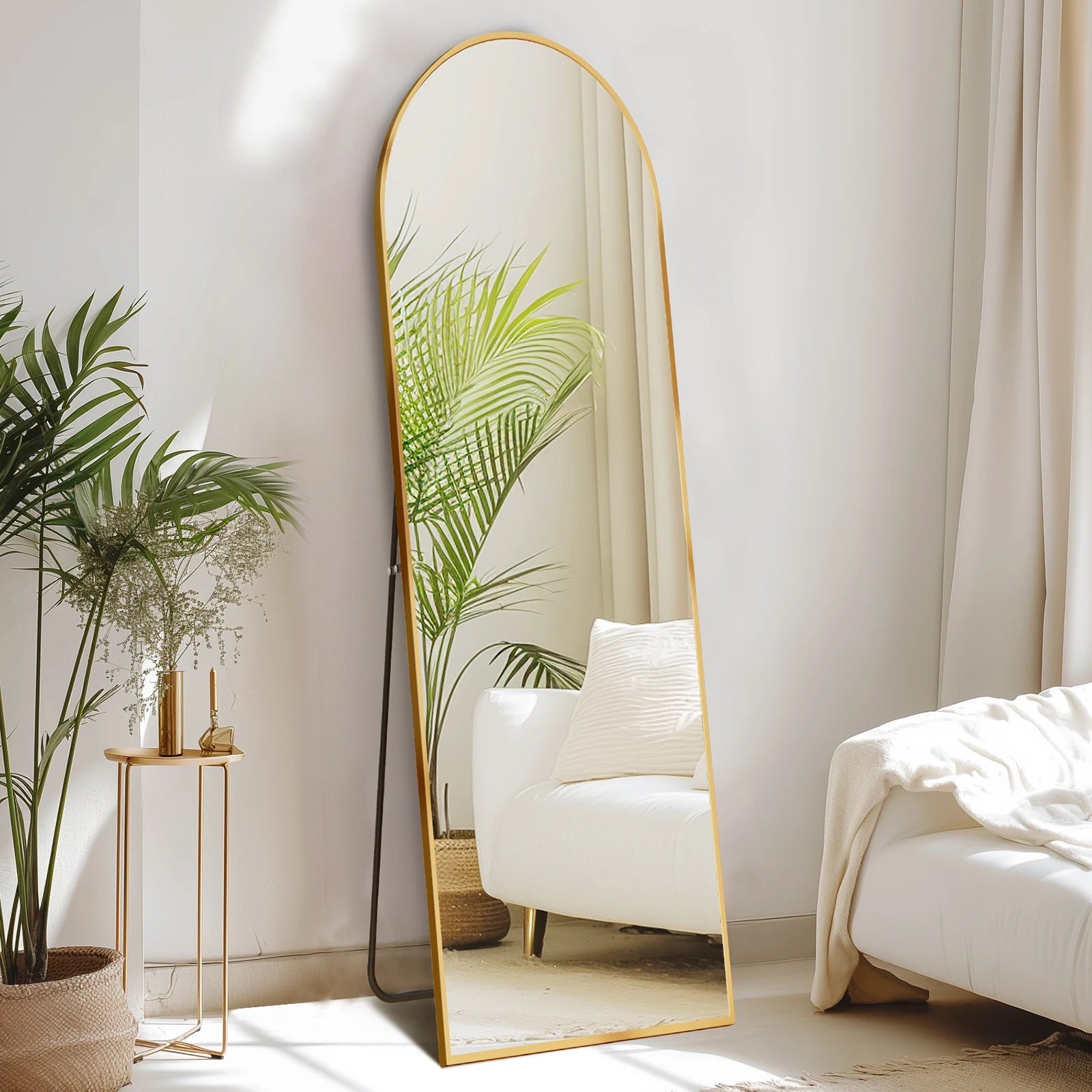 Arched full length floor mirror with gold metal frame in modern minimalist bedroom with white chair and plants