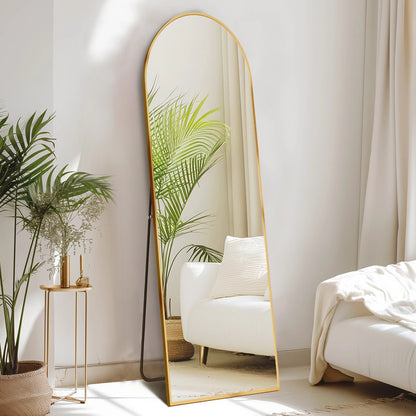 Arched full length floor mirror with gold metal frame in modern minimalist bedroom with white chair and plants