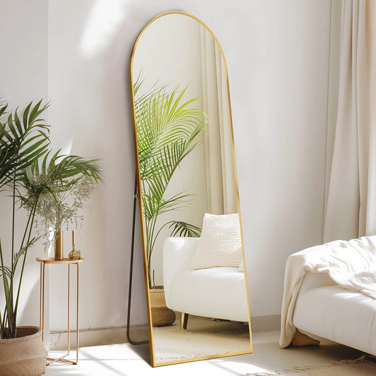 Arched full length floor mirror with gold metal frame in modern minimalist bedroom with white chair and plants
