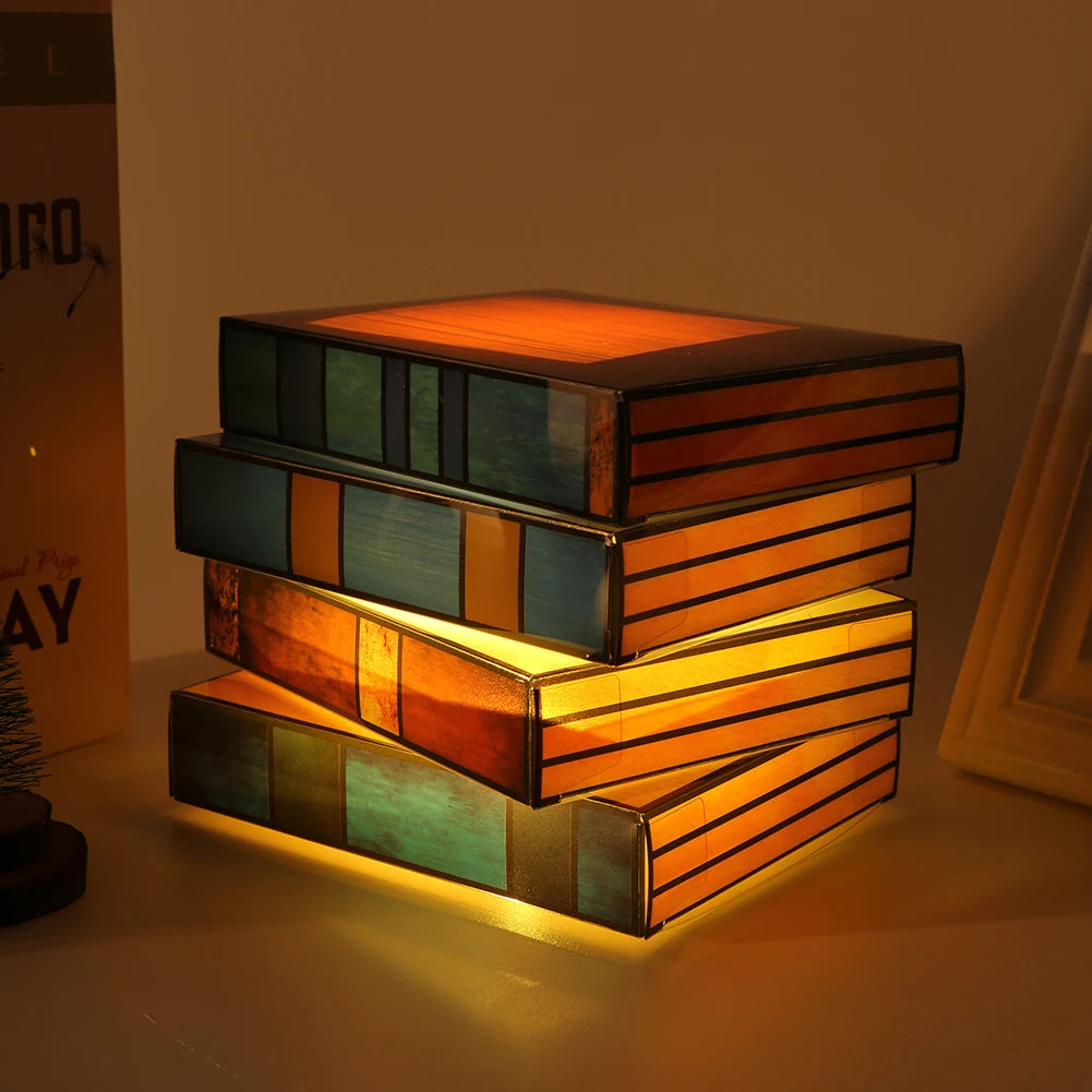 Stained glass lamp shaped like stacked books glowing warm amber and teal, displayed on a desk as decorative night light.