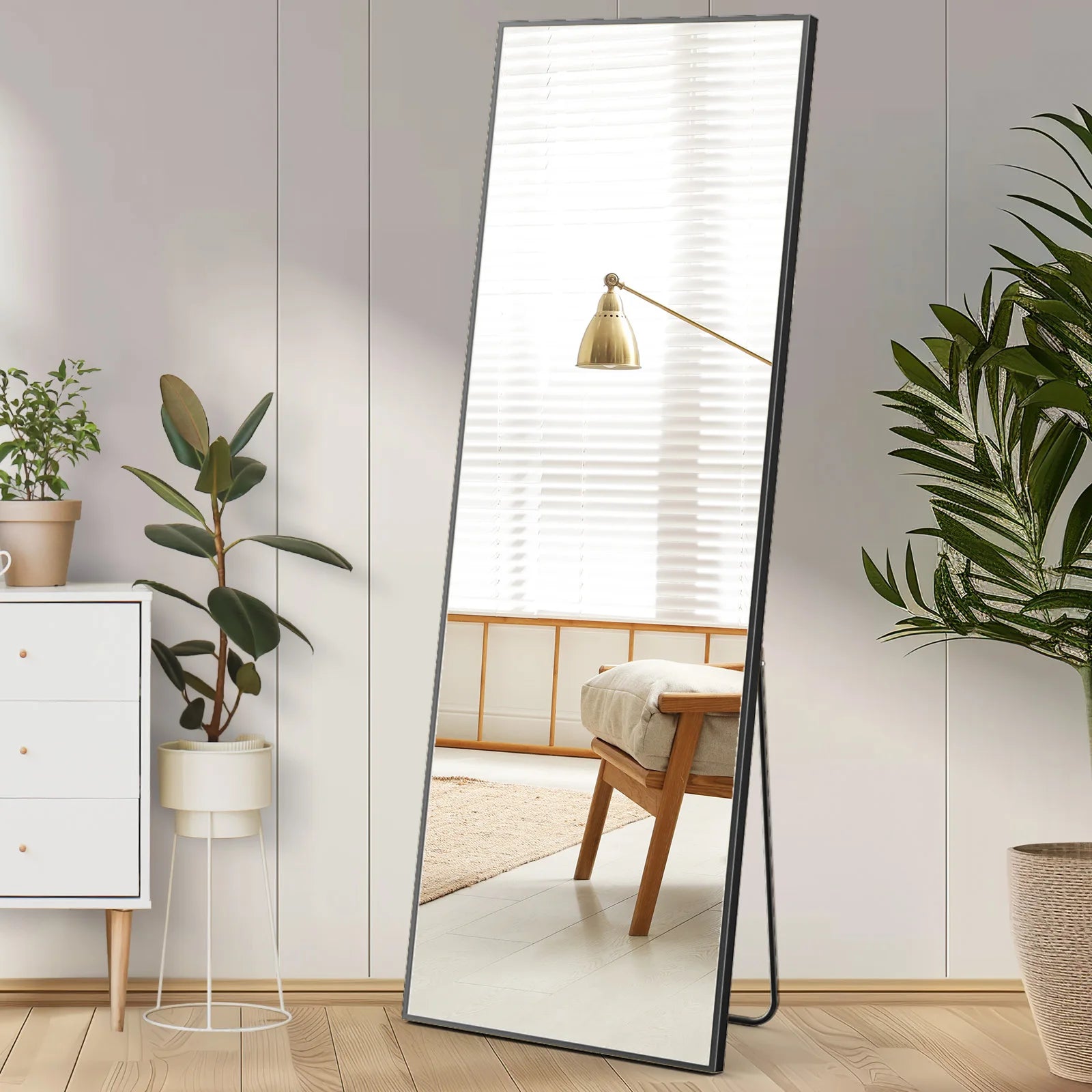 Full-length rectangular standing mirror with black metal frame leaning against white wall in modern bedroom setting.