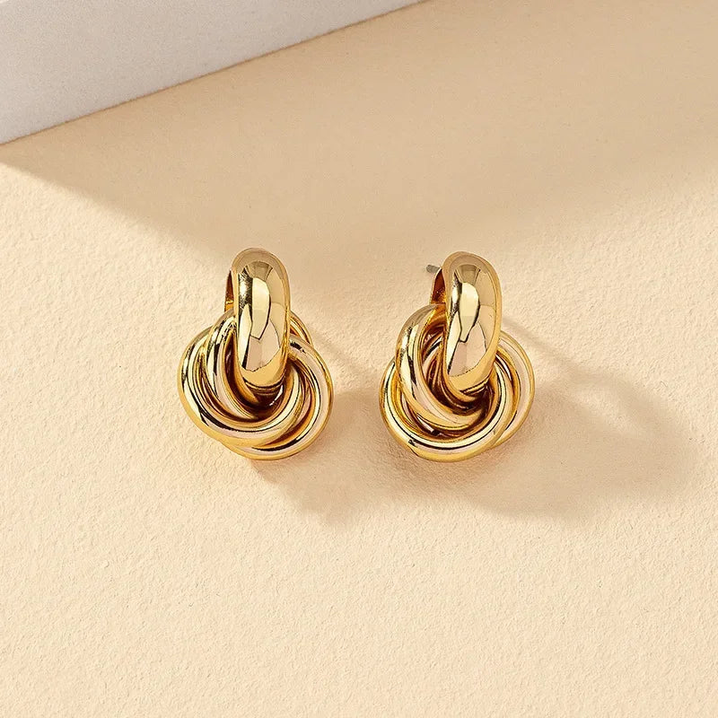 Gold tone interlocked knot hoop earrings for women on beige background, chunky twisted metal design statement jewelry