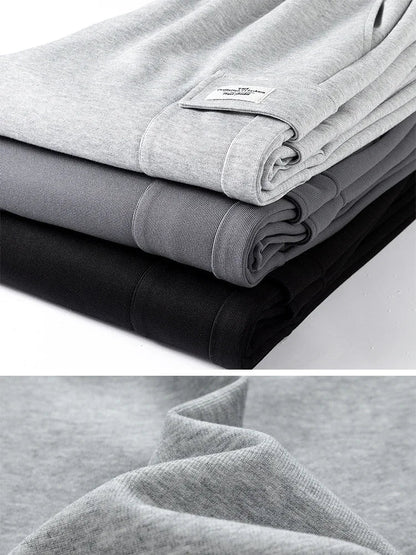 Three folded men's relaxed fit sweatpants stacked in light gray, dark gray, and black with close-up fabric detail.
