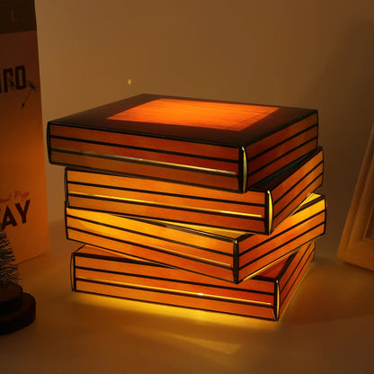 Glowing stained glass lamp shaped like stacked books casting warm amber light on a desk surface.
